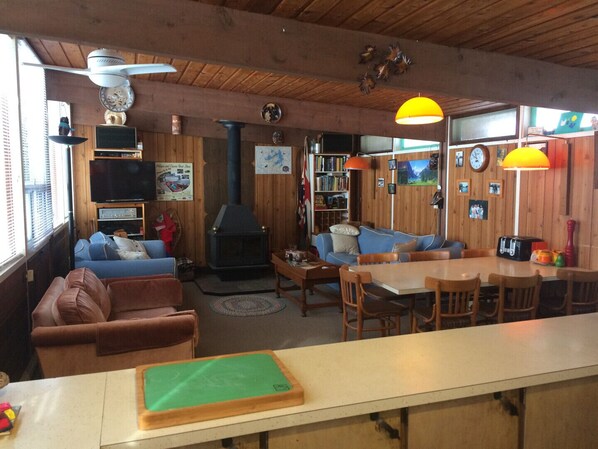 A cozy cabin interior with a fireplace, wooden walls, a sofa, a dining table, and a bookshelf.