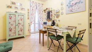 A dining area with a table set for two, a green folding chair, a cabinet with floral patterns, and a window with floral curtains. - Dreaming of You Bed & Breakfast (Assisi)