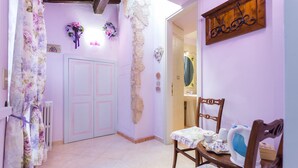 A hallway with a white door, floral curtains, a wooden chair, and a table with a teapot. - Dreaming of You Bed & Breakfast (Assisi)