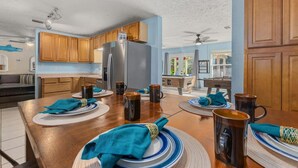 A spacious kitchen with wooden cabinets, a stainless steel refrigerator, and a dining area set for six with blue and white plates and napkins. - Pirates Cove ~ Heated Pool, Hot Tub, Game Room & 6-Seater Golf Cart (Panama City Beach)