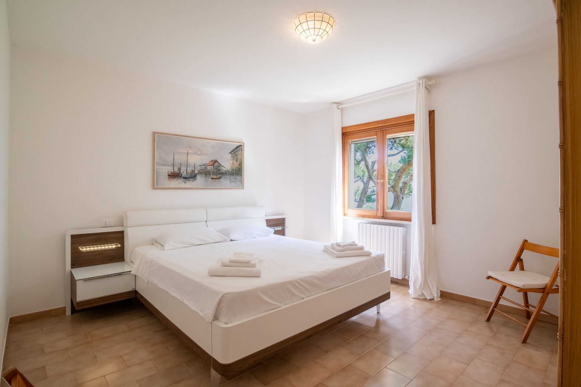 A bedroom with a bed, a nightstand, a chair, a window with curtains, and a painting on the wall.