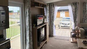 Un espace de vie compact avec un téléviseur posé sur un meuble en bois, une petite table sur laquelle se trouvent un globe terrestre et un bol, et un balcon donnant sur une voiture et une clôture blanche.