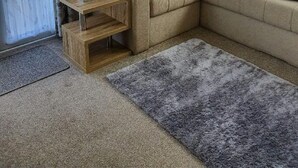 Un salon avec un canapé d’angle beige, une petite table d’appoint en bois et un tapis à poils longs gris.