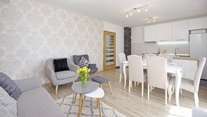 A modern living room with a grey sofa, a small round table with a vase of flowers, and a dining area with white chairs and a table.