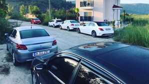 Ein zweistöckiges Haus mit Balkon, geparkten Autos und ländlicher Umgebung im Hintergrund.