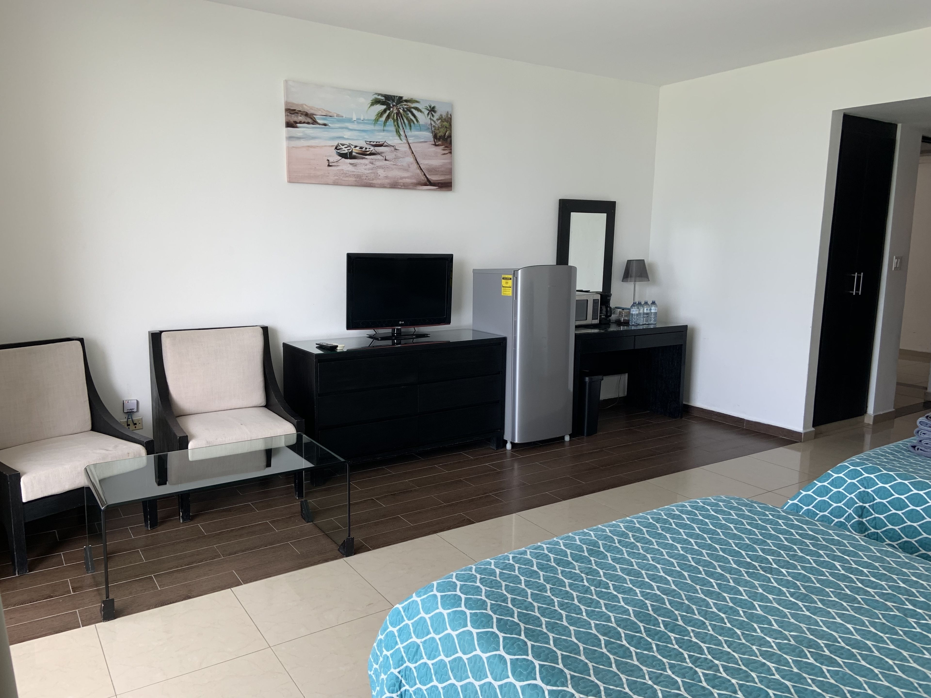 Habitación de hotel con cama, escritorio con televisor, refrigerador y un cuadro con una playa.