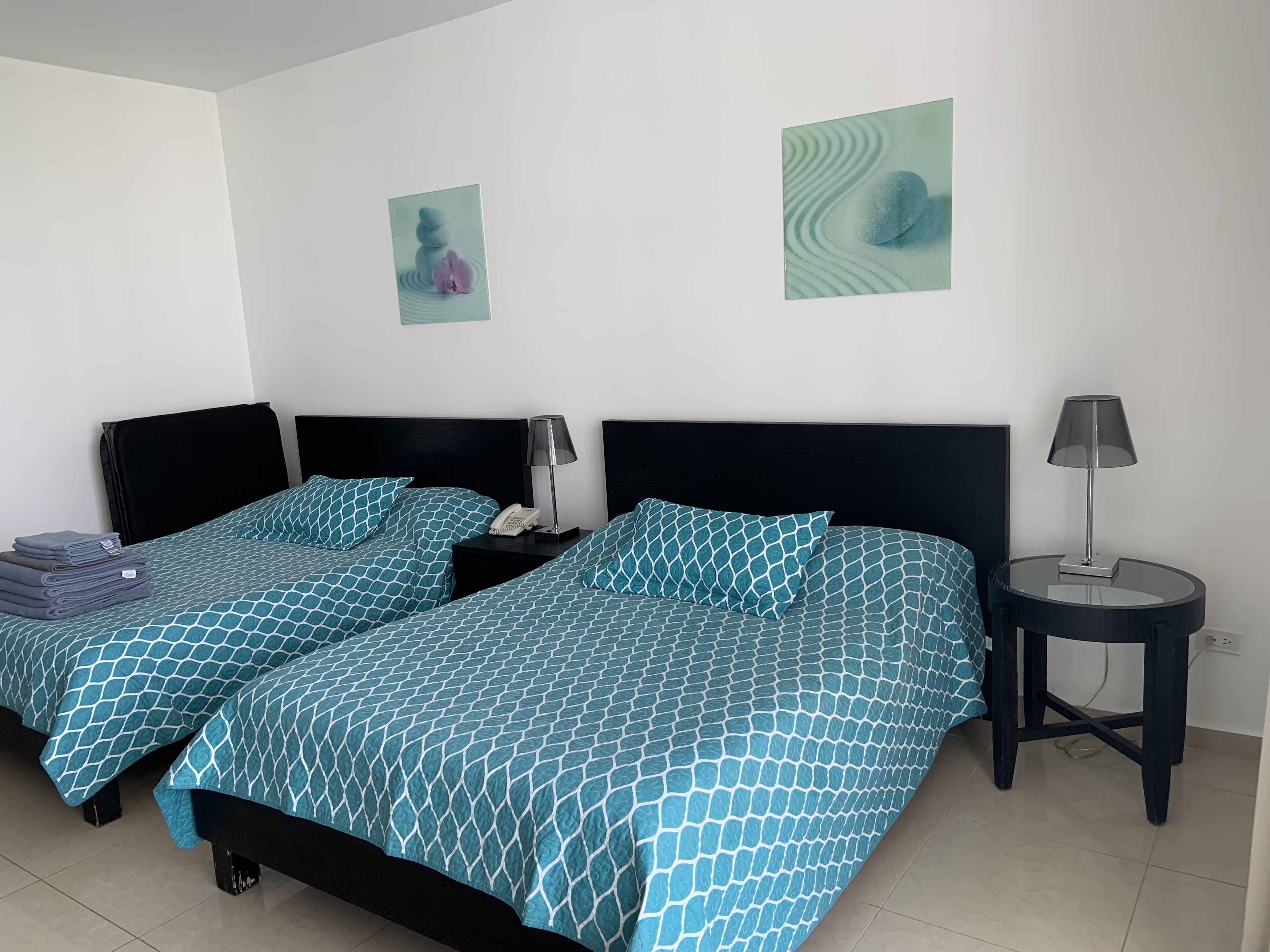 A hotel room with two beds, each with a black headboard and a blue patterned bedspread. There are two bedside tables, one with a lamp and the other with a phone. The walls are white, and there are two framed pictures on the wall.