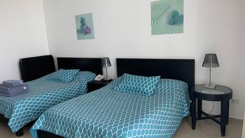 A hotel room with two beds, each with a black headboard and a blue patterned bedspread. There are two bedside tables, one with a lamp and the other with a phone. The walls are white, and there are two framed pictures on the wall.