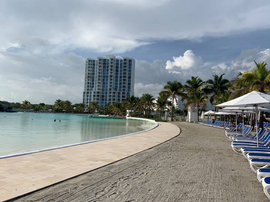 A beachside pool area with lounge chairs and umbrellas, palm trees, and a tall building in the background.