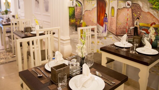 Comedor con mesas dispuestas para una comida, con sillas blancas, mesas de madera oscura y un mural que representa una escena callejera al fondo.