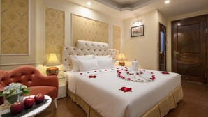 Una habitación de hotel con una cama grande, un sillón rojo, una mesita con manzanas y flores, y una puerta de madera.