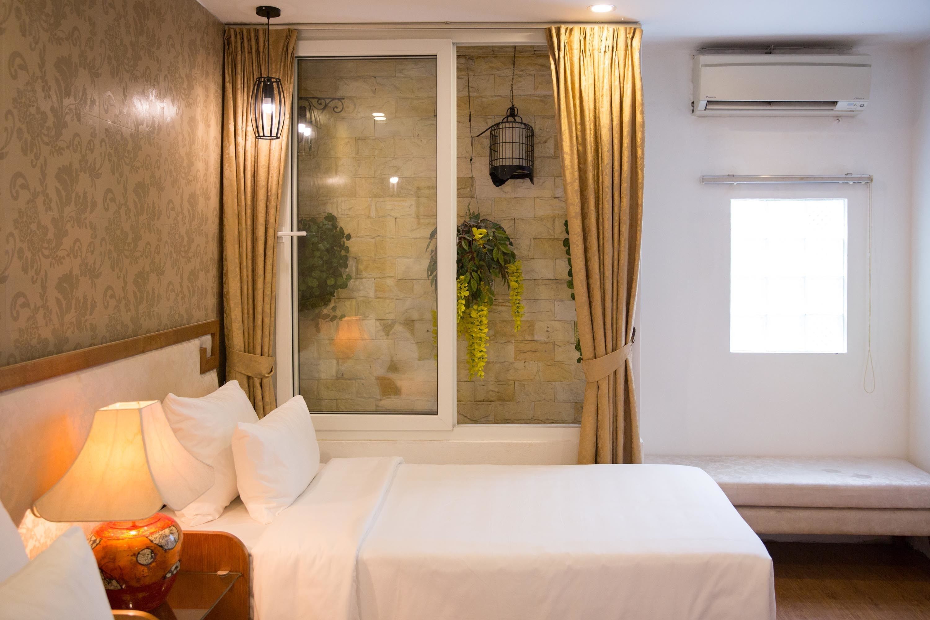 Quarto de hotel com uma cama, mesa de cabeceira com luminária, janela com cortinas, ar condicionado fixado na parede e uma pequena varanda com uma planta.