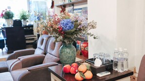 Un comedor con una mesa dispuesta, adornada con un florero, frutas y un control remoto.
