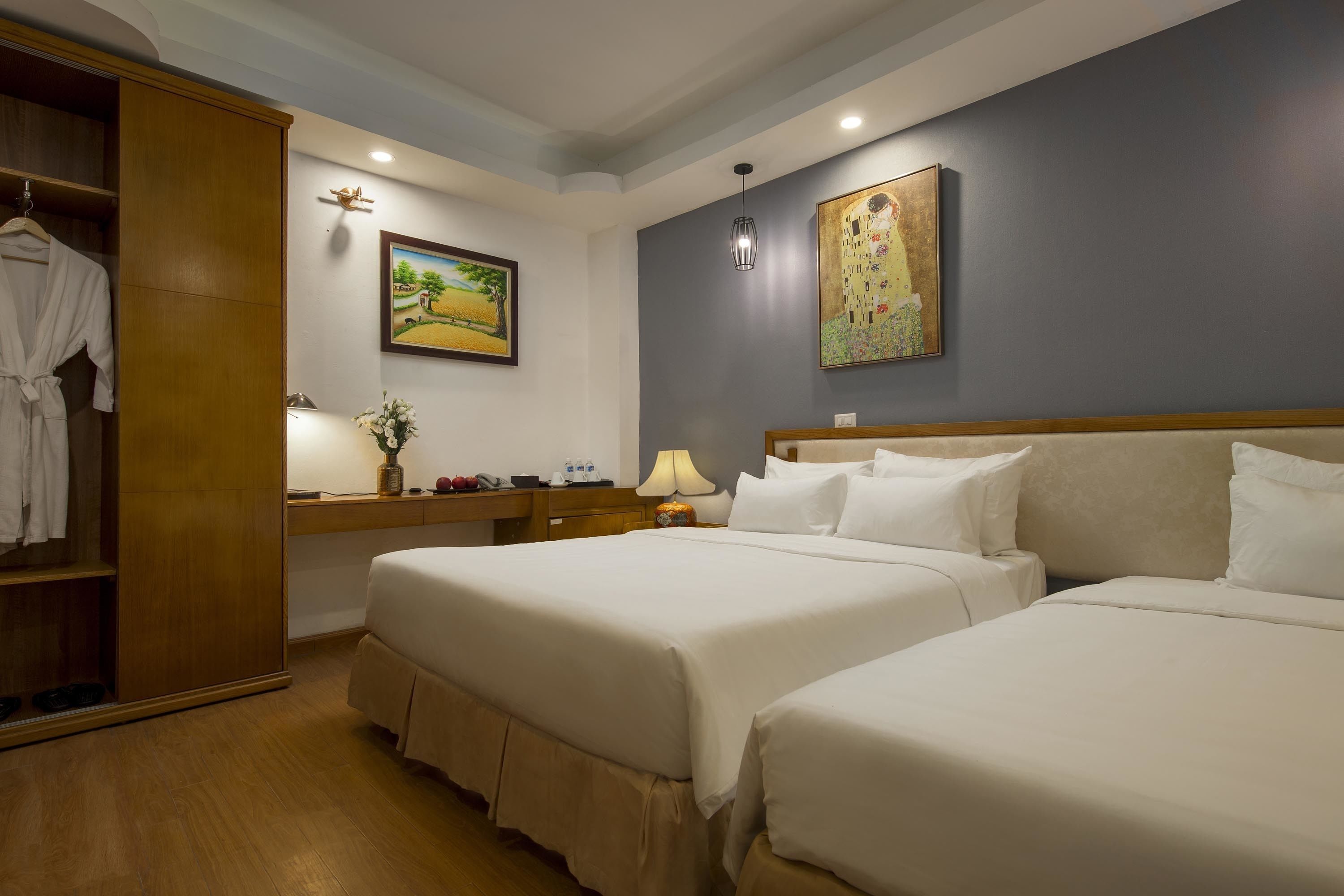 A hotel room with two beds, a wooden wardrobe, a desk with a lamp, and artwork on the walls.