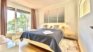 A modern bedroom with a large bed, a desk, and a view of trees through the window. - Nautilus-Bay Suites - Apt. 420 Three-room suite for 2-6 persons (Zavala)