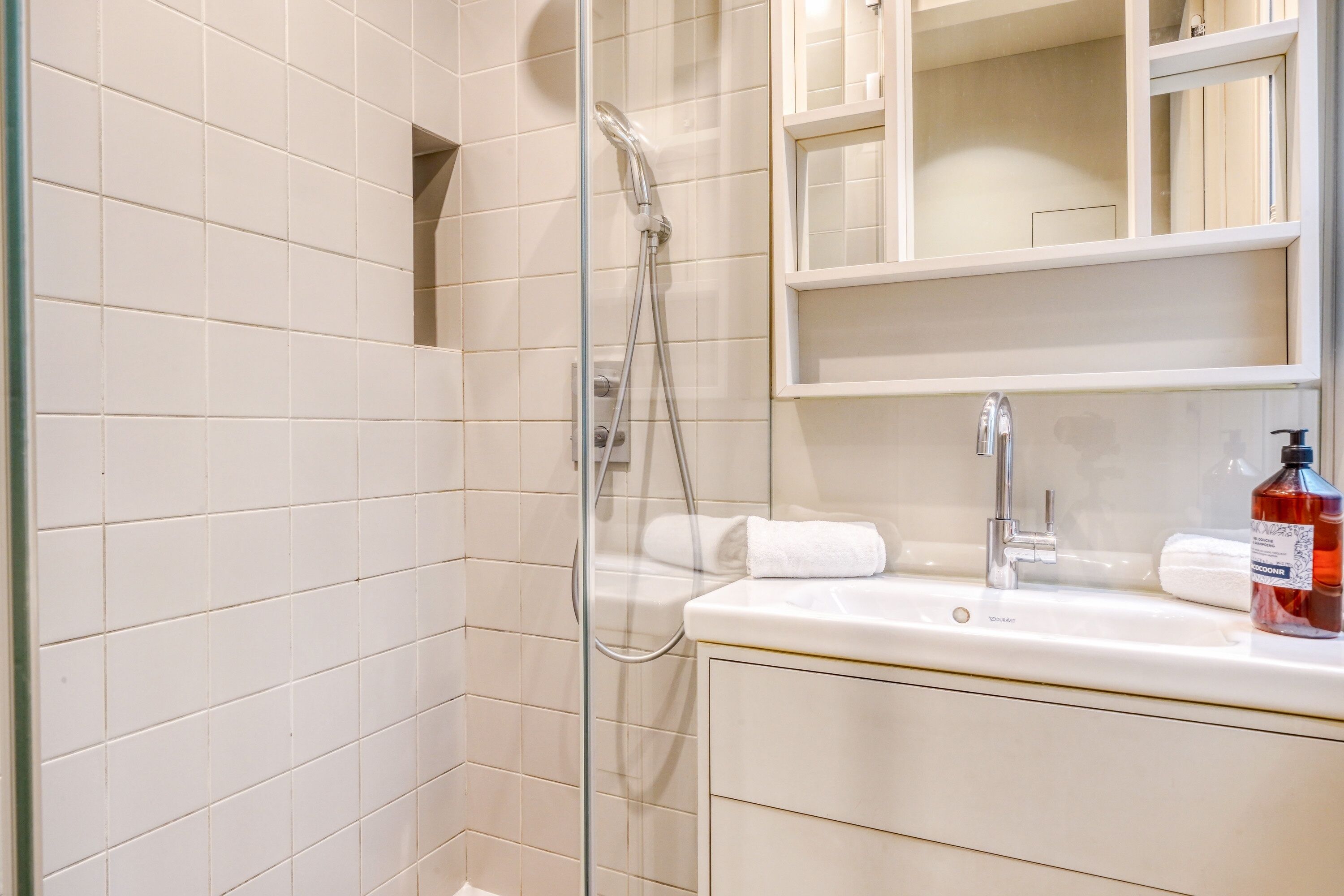 Une salle de bain avec une douche à parois de verre, un lavabo avec un miroir au-dessus et un flacon de savon liquide posé sur le lavabo.