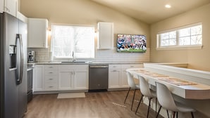 A modern kitchen with white cabinets, a stainless steel refrigerator, a dishwasher, and a dining area with a long table and four chairs.