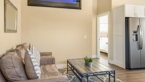 A living room with a sofa, a coffee table with a plant, a framed picture on the wall, and a kitchen area with a refrigerator.