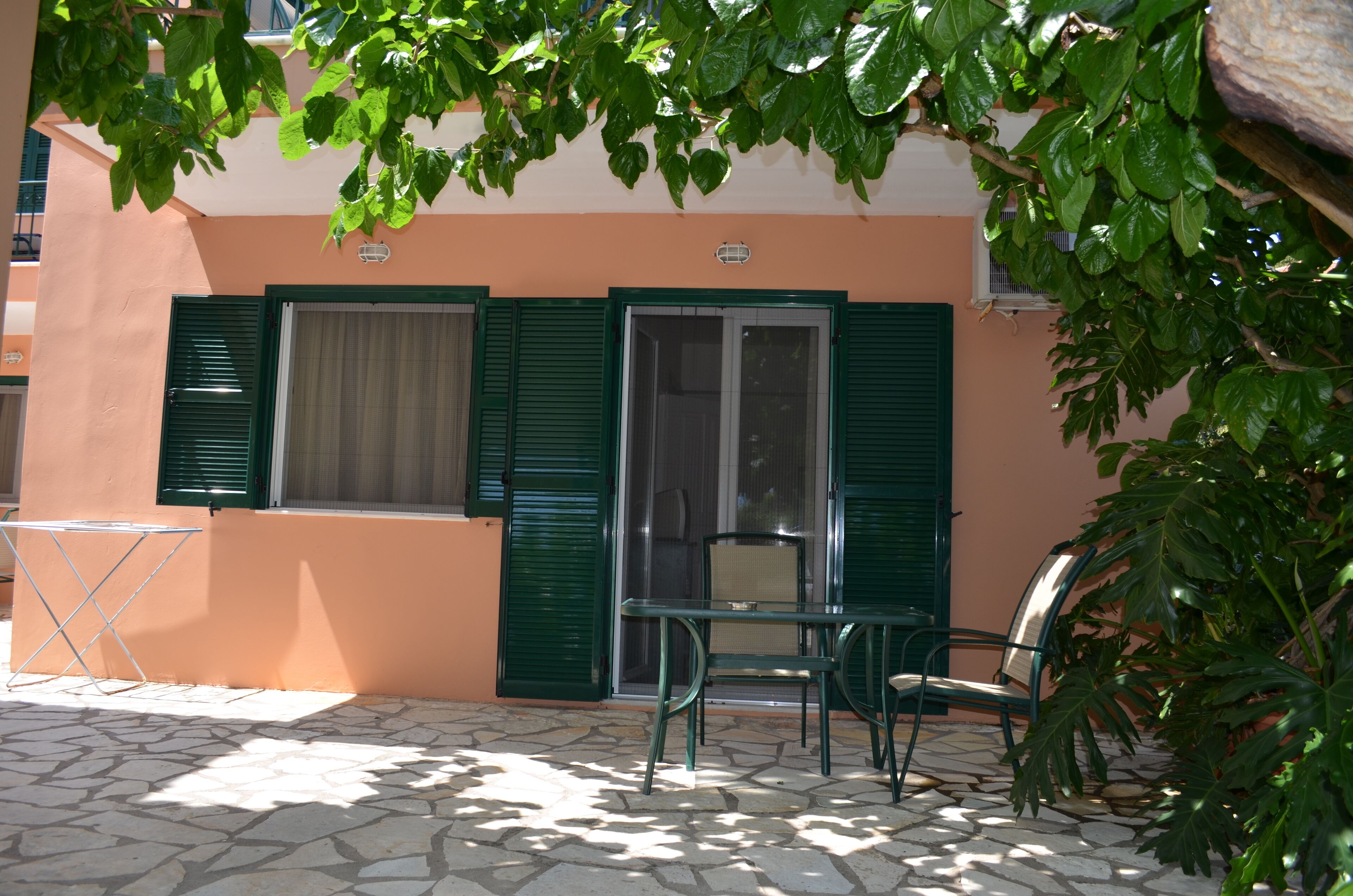 Un patio con una mesa, sillas y una silla plegable, y una puerta con contraventanas verdes.