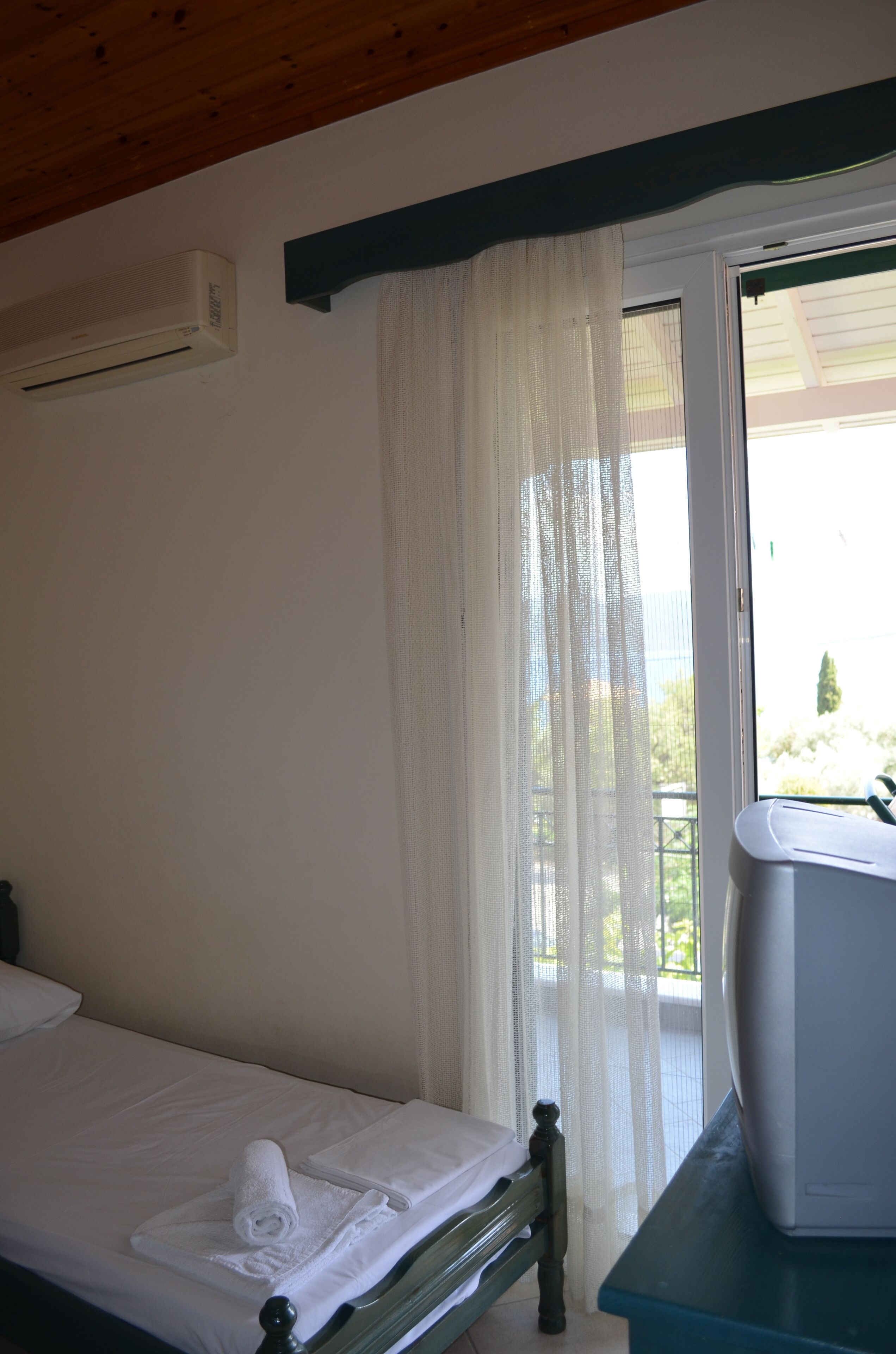 Ein Schlafzimmer mit einem Bett, einem Fenster mit durchsichtigen Vorhängen, einer weißen Klimaanlage und einem kleinen Tisch mit einem weißen Gerät.