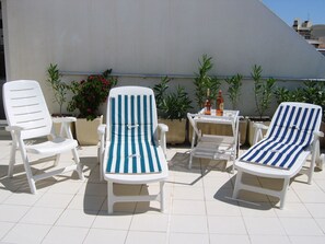 Zwei weiße Lounge-Stühle mit blauen und weißen Streifen, ein kleiner Tisch mit Flasche und Glas, und Topfpflanzen vor einer weißen Wand.