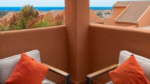 Een balkon met twee witte stoelen met kussens en oranje kussens, met uitzicht op een kustlandschap met gebouwen en bomen.
