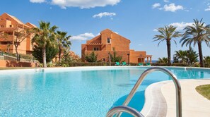 A swimming pool with clear blue water, surrounded by palm trees and a building with a flat roof. - Luxury Apartment On Santa Maria Golf And Country Club (Marbella)
