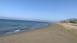 Een strand met een heldere lucht, een rustige zee en verre bergen.