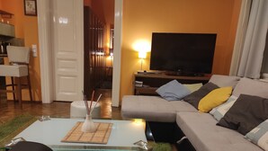 Een woonkamer met een glazen salontafel, een bank met kussens, een flatscreen televisie en een eethoek met een stoel en tafel.