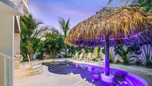 A pool area with a thatched umbrella, lounge chairs, and tropical plants. - Beach Views - Family-Friendly, Steps to Lido Beach! (Lido Key)