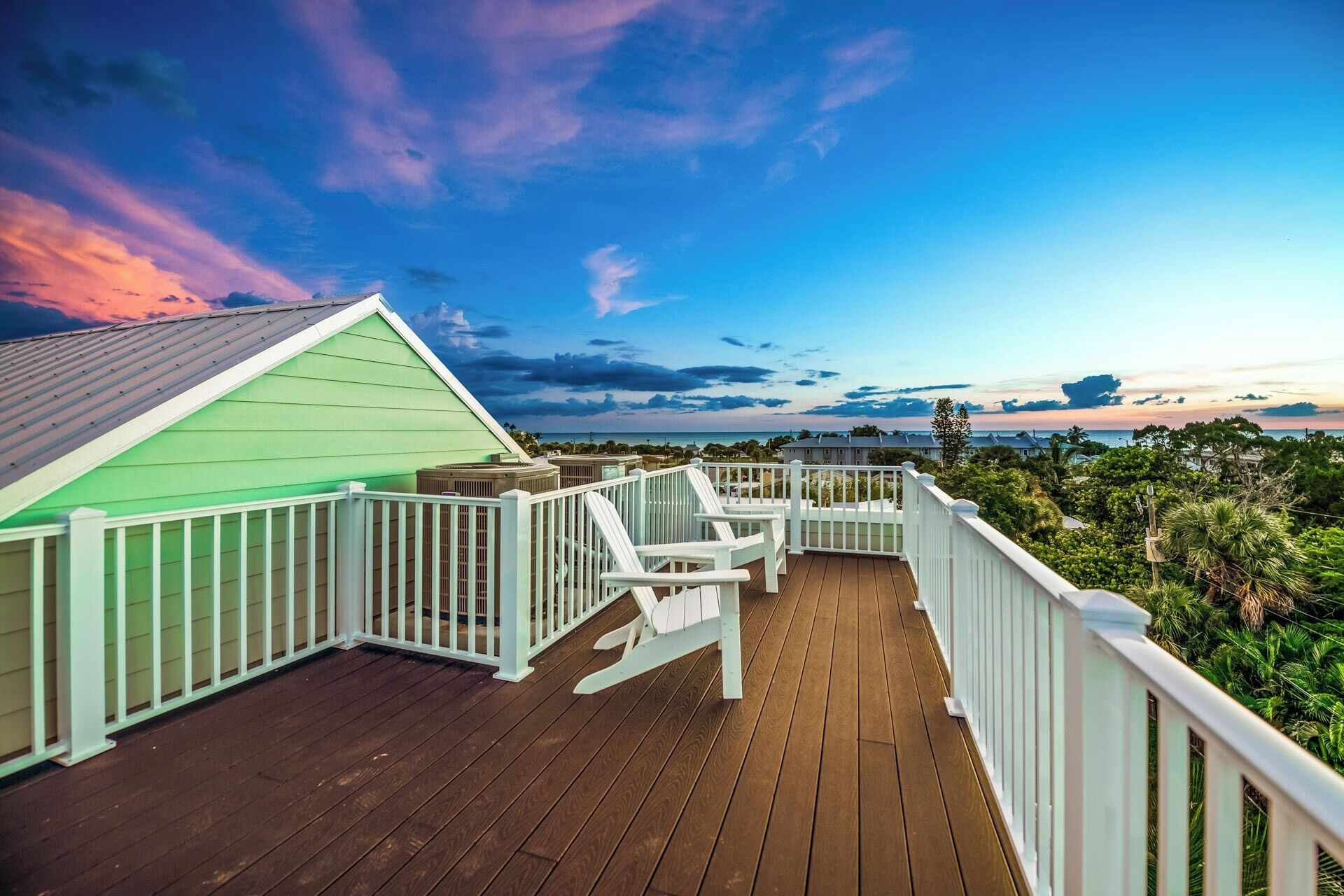 A wooden deck with white railings, a green building, and a view of the ocean and sky.