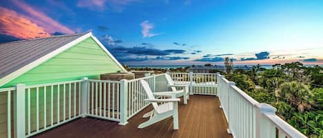 A wooden deck with white railings, a green building, and a view of the ocean and sky.