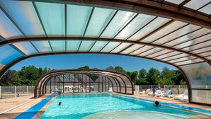 A covered swimming pool with clear blue water, a curved roof, and a view of a house in the background. - Studio proche de la Plage! (Auberville)