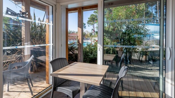 A covered patio with a table and chairs, large glass doors, and a view of a swimming pool and trees. - Seulement à 400m du lac de Biscarrosse | Piscines! (Parentis-en-Born)