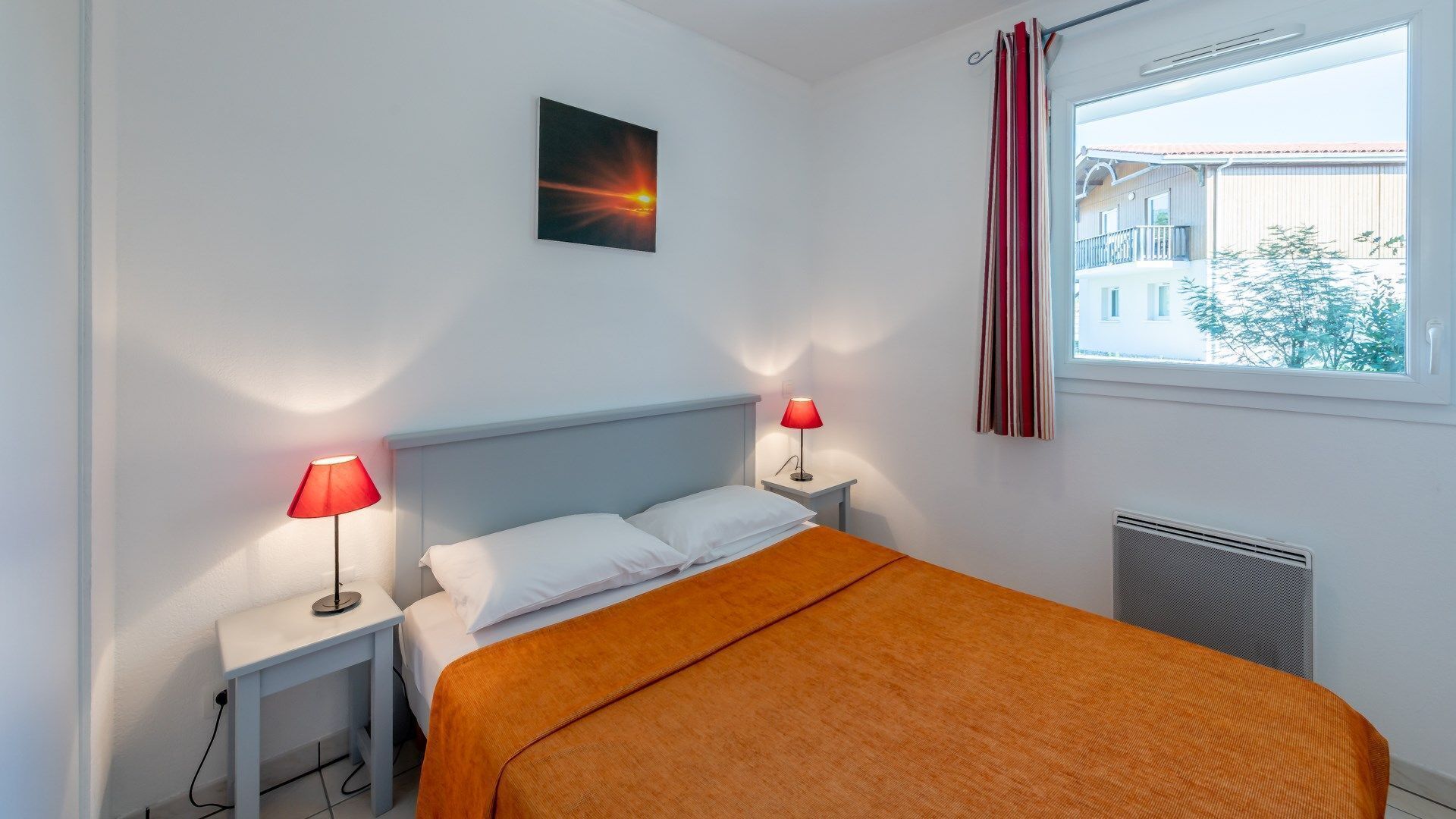 Ein Schlafzimmer mit einem Bett, zwei Nachttischen mit Lampen, einem Fenster mit Vorhängen und einem Bild an der Wand.