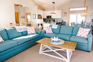 A spacious living room with teal sofas, a wooden coffee table, and a kitchen in the background. - ☀️Steps to Beach, Private Pool & Hot Tub☀️ (Nags Head)