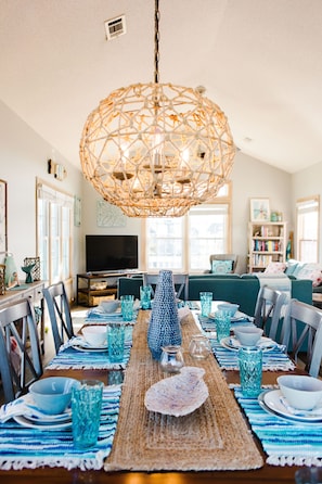 A dining area with a central table set for four, surrounded by chairs. A large woven chandelier hangs above, and a bookshelf is visible in the background. - ☀️Steps to Beach, Private Pool & Hot Tub☀️ (Nags Head)