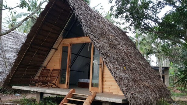 Una cabaña con techo de paja, puerta de madera y un pequeño porche con dos sillas de madera.