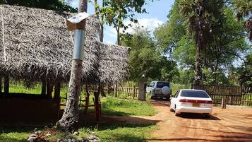 A rural road with a thatched-roof hut, a tree, and a parked car.