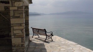 A stone bench by a waterfront with a stone building in the background. - Zmas Studios (South Pelion)