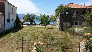 A stone building with a tiled roof, a fenced yard with yellow flowers, and a view of the sea and mountains in the background. - Zmas Studios (South Pelion)