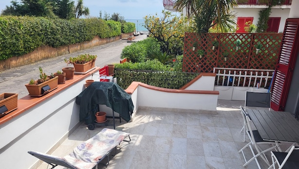 A terrace with a table and chairs, a covered grill, potted plants, and a view of the sea. - Formia Seaside Apartment With Beautiful Terrace (Formia)