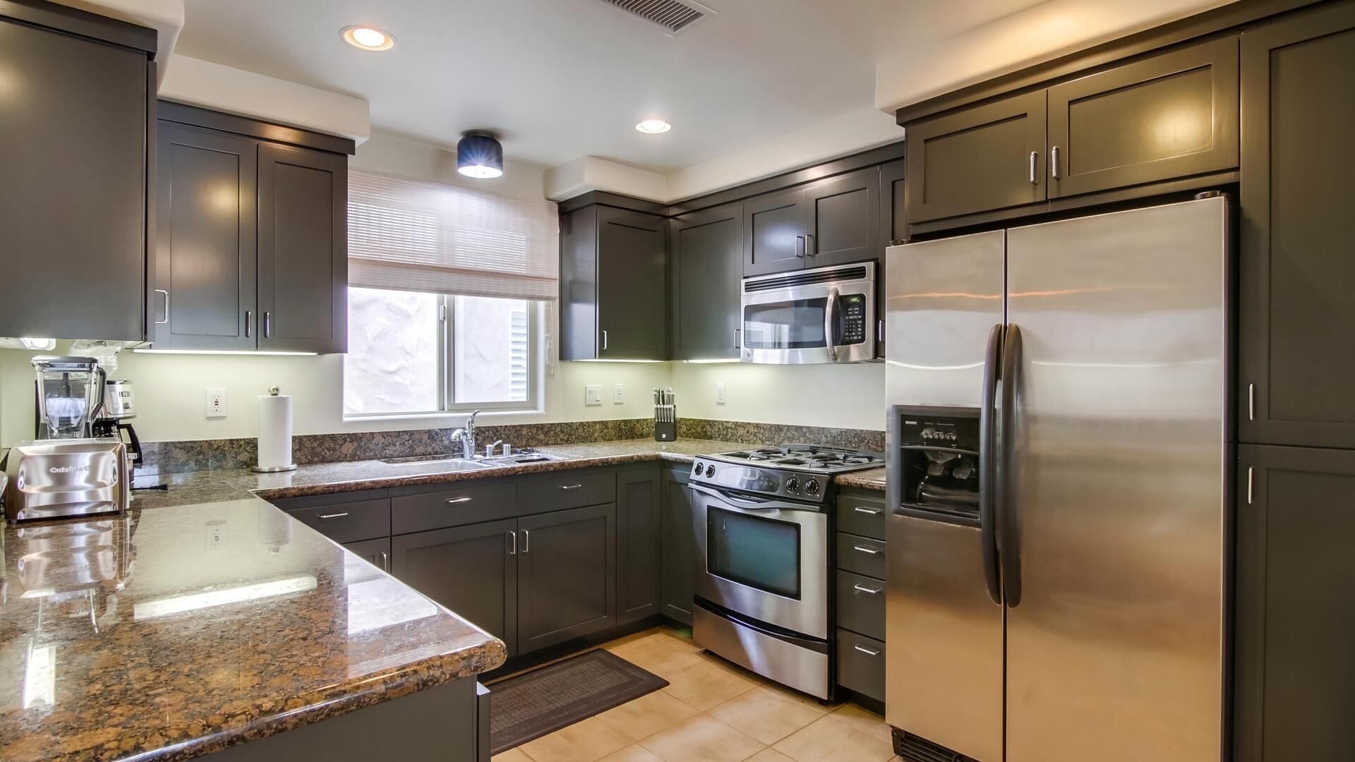 A modern kitchen with dark cabinets, stainless steel appliances, granite countertops, and a window with blinds.