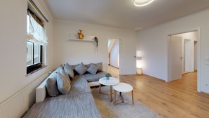A modern living room with a grey sectional sofa, a round white coffee table, and a small side table. There is a window with sheer curtains, a white radiator, and a plant on a shelf. The room has wooden flooring and a hallway leading to another room. - Apartment in historical building - old school (Spittelstein)
