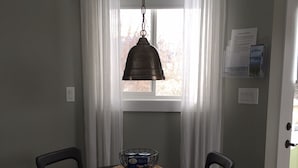 Un coin repas comprenant une table ronde, quatre chaises, un luminaire suspendu et une fenêtre avec des rideaux.