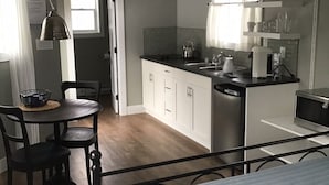 Une cuisine compacte avec des meubles blancs, un évier et une machine à café. On y trouve une table à manger avec des chaises et un luminaire suspendu.