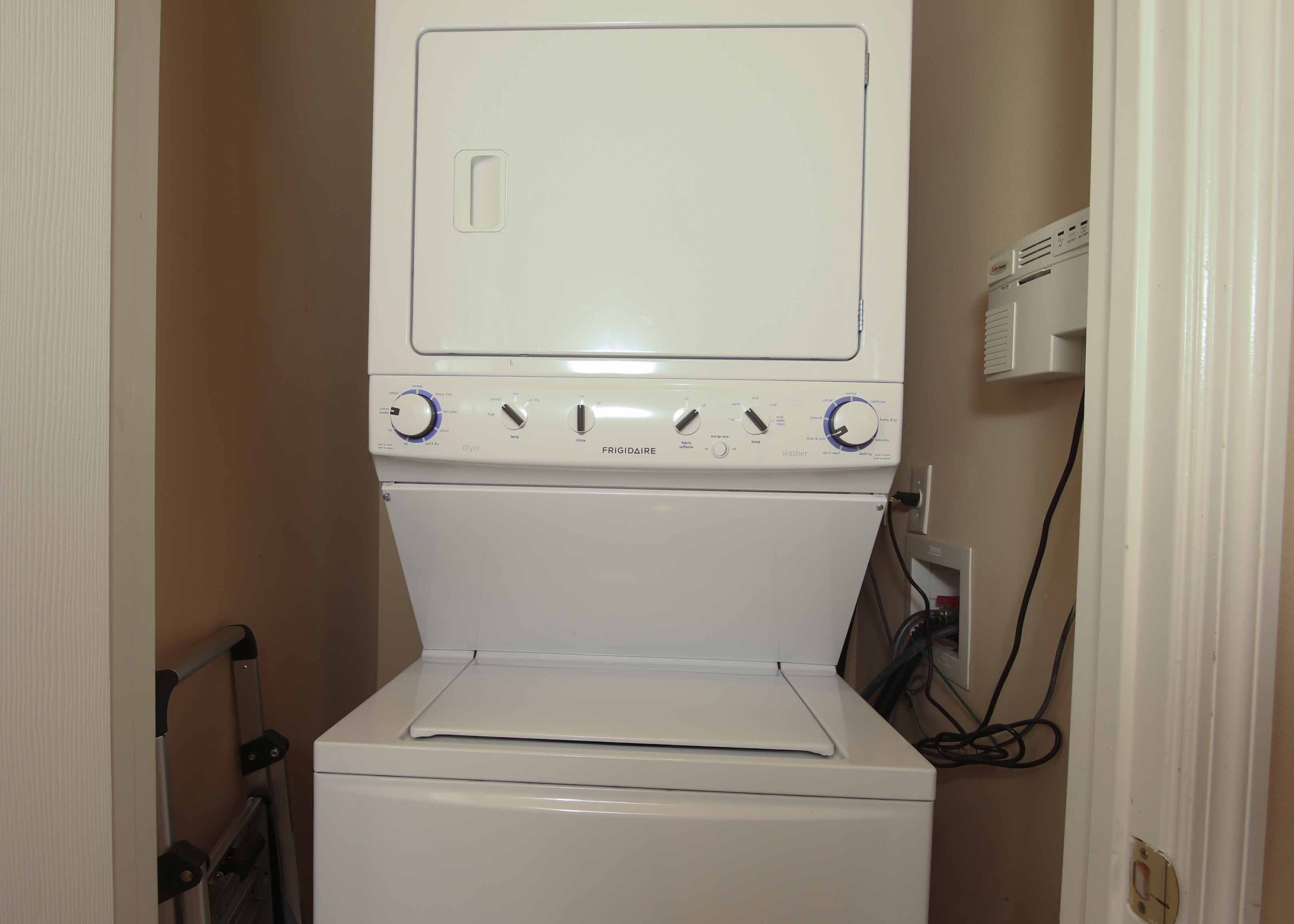 A compact, white, two-door washer and dryer unit with control knobs and a front-loading door, situated in a narrow hallway with beige walls.