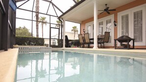 Piscina com fundo de vidro, rodeada por cadeiras e uma mesa, sob uma área coberta com ventoinha no teto.