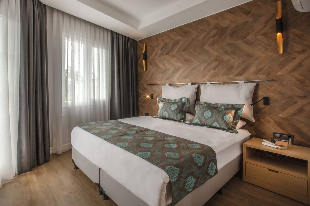 Una habitación de hotel moderna con una cama grande, paredes de madera con un patrón de espiga, una mesita de noche con lámpara y cortinas translúcidas en la ventana.