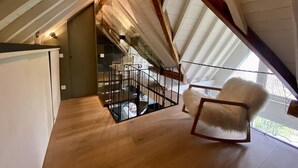 Een slaapkamer in loftstijl met een schuine kap, houten balken, een glazen trap en een pluizige witte fauteuil.